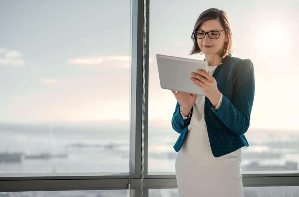 A woman in a blue jacket and glasses using a tablet near a large window with a city view.