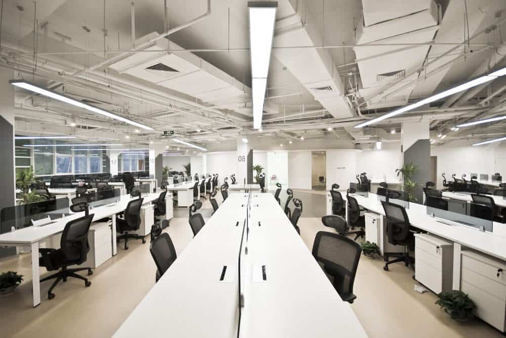 Modern open office space with rows of desks and chairs, large ceiling lights, and minimal decor.
