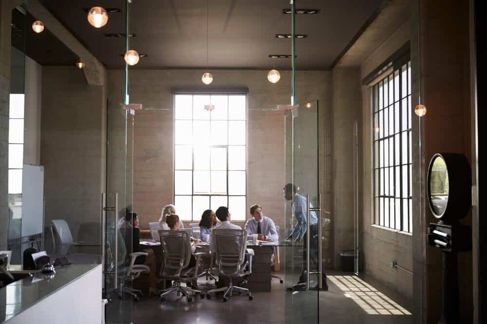 People having a meeting around a table in a modern conference room with large windows and hanging lights.