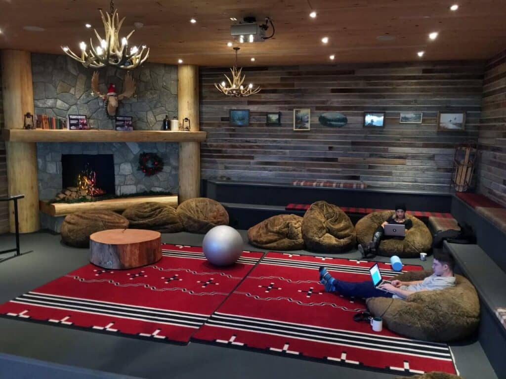 Cozy lounge with bean bags, red rugs, a stone fireplace, and two people using laptops.
