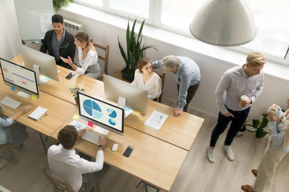 Office with people working at desks, discussing and viewing computer screens with charts. Some hold coffee cups.