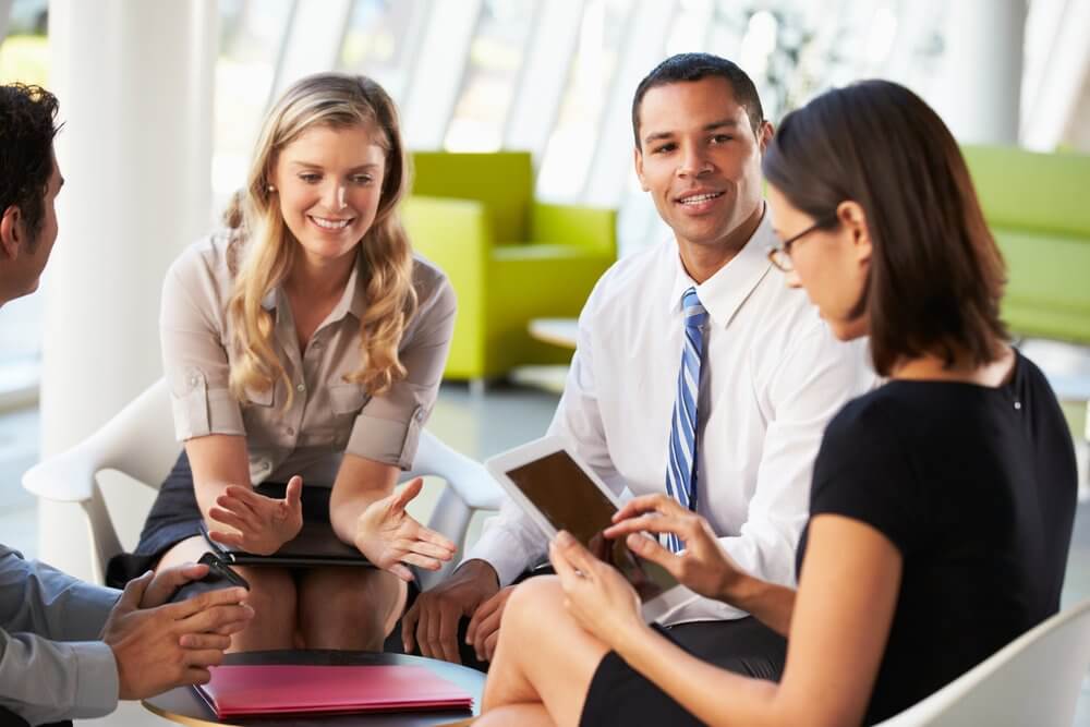 Four people in business attire have a discussion around a table in a bright office setting.