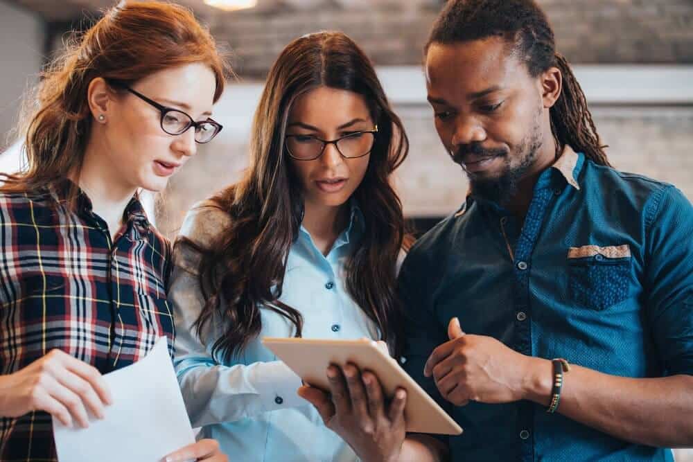 Three people collaborating, looking at a tablet. One holds a paper, all appear focused and engaged.