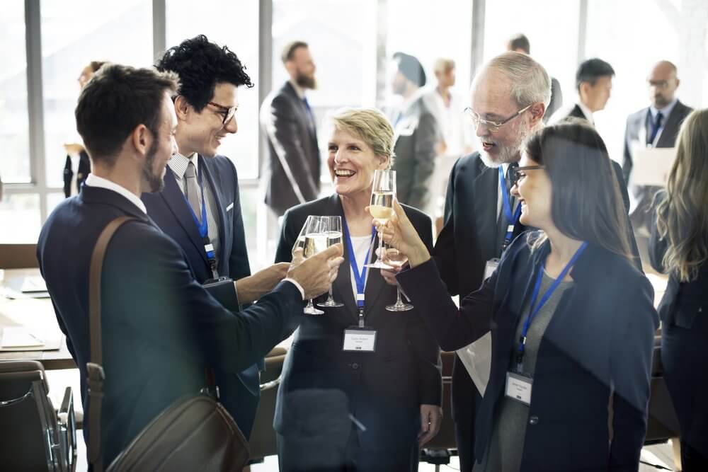 Group of business professionals clinking glasses and smiling at a networking event in a bright conference room.