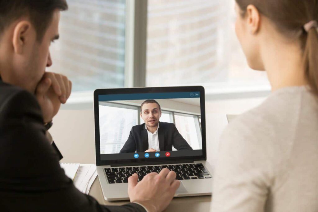 Two people having a video call with a man on a laptop screen in a bright office setting.