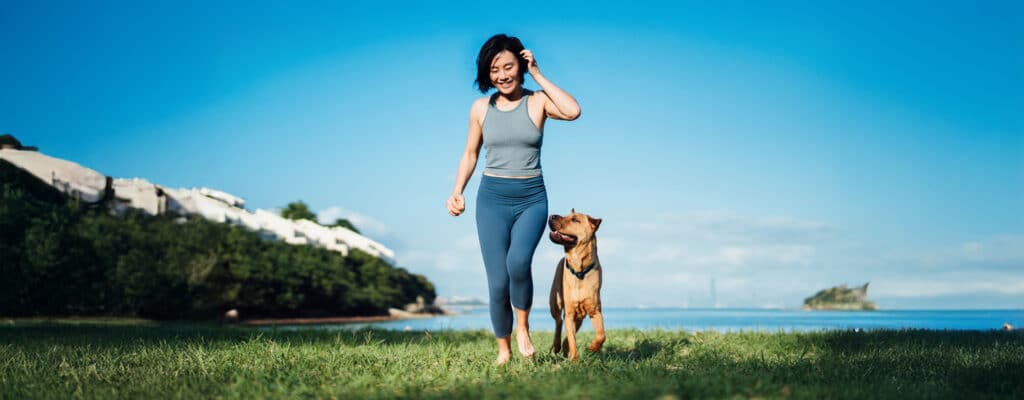Woman jogging with a dog on a grassy field near water, clear blue sky above.