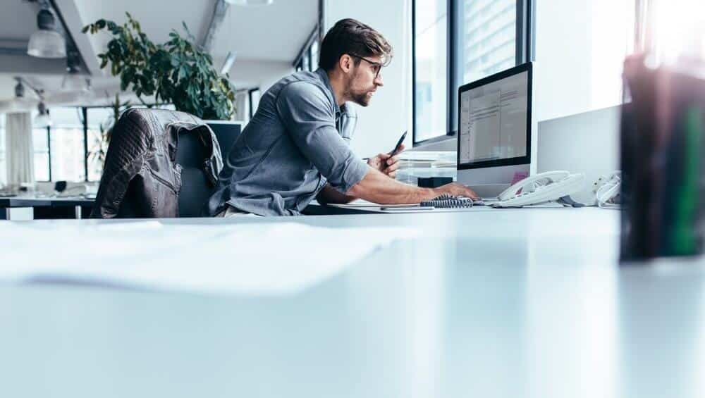 Man sitting at a desk in an office, using a computer and holding a pen, with plants in the background.