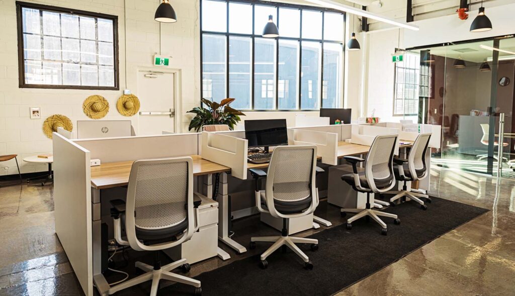 A modern office with six workstations, white chairs, and large windows letting in natural light.