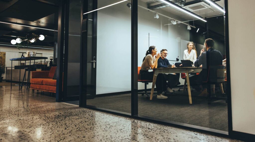 People sitting around a conference table in a modern office, viewed through a glass wall.
