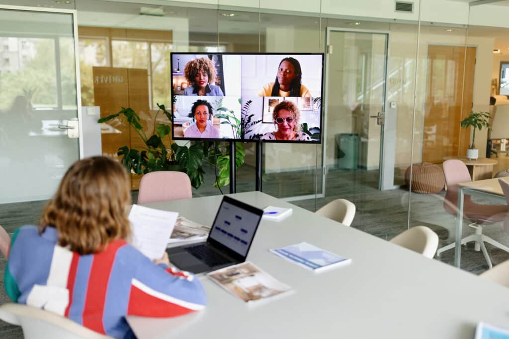 Person in a colorful sweater at a conference table, video conferencing on a large screen in a modern office.