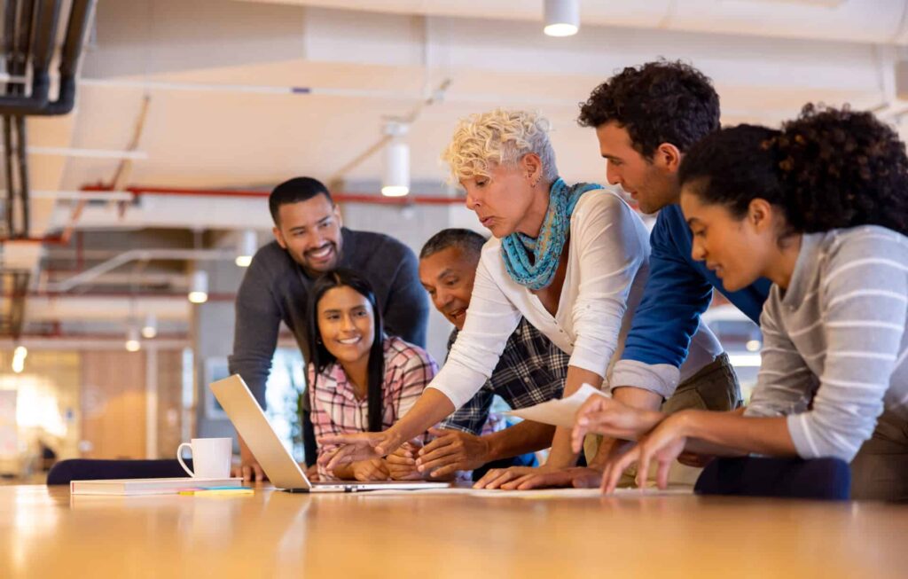 Diverse group of people gathered around a laptop in a modern office setting, focused and collaborating.