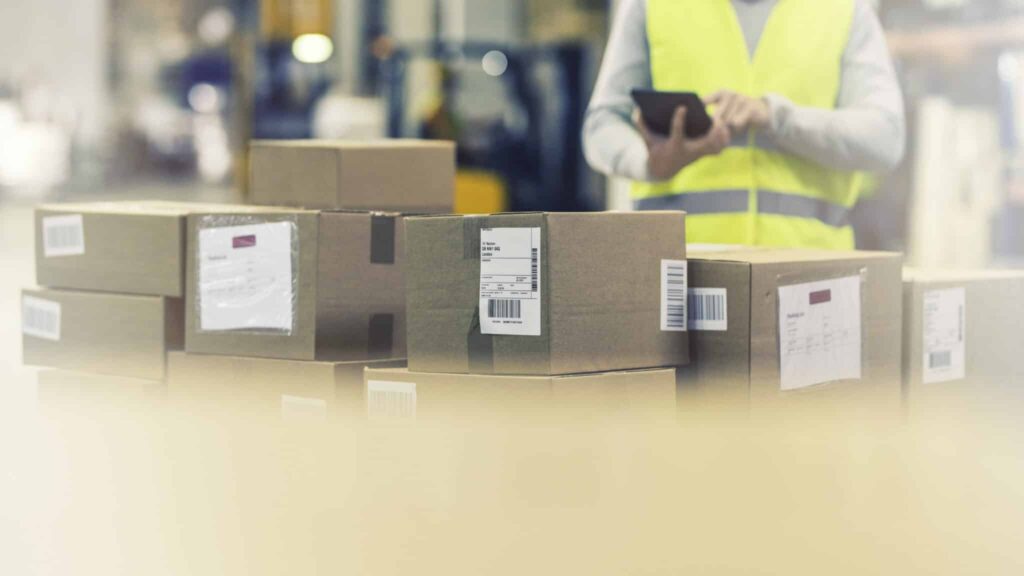 Person in yellow vest holds a tablet near stacked cardboard boxes with labels in a warehouse.
