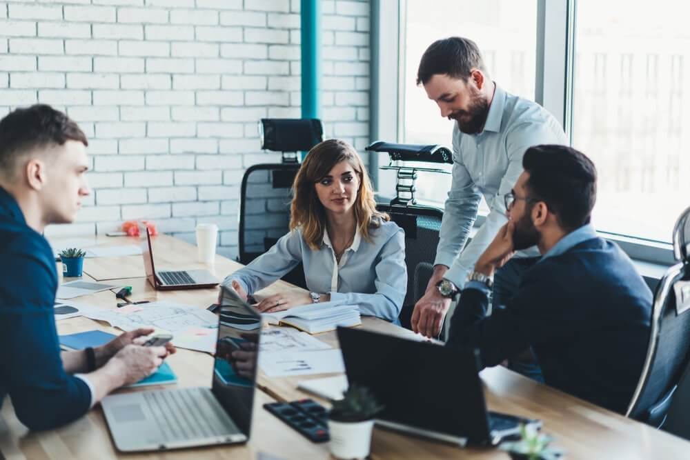 Four people having a discussion around a table with laptops and papers in a modern office setting.