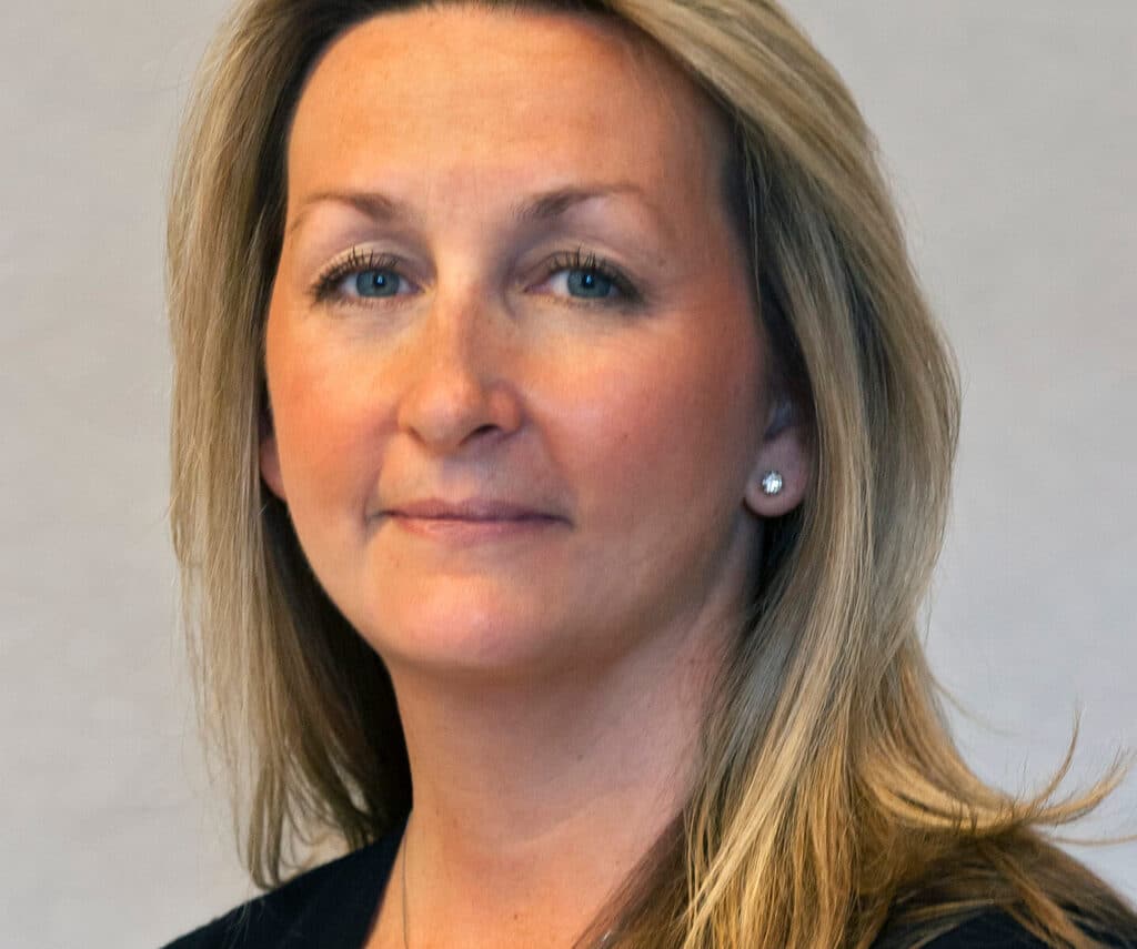 A woman with long blonde hair and subtle jewelry, wearing a dark top, looking directly at the camera.