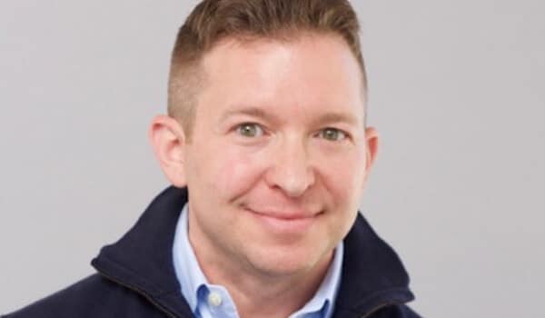 Smiling man with short hair wearing a blue collared shirt and dark jacket against a gray background.