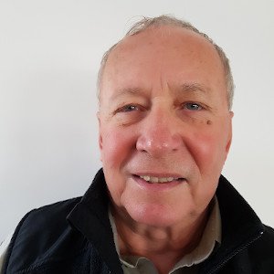 Elderly man with light skin, short gray hair, wearing a black jacket over a beige shirt, smiling slightly.