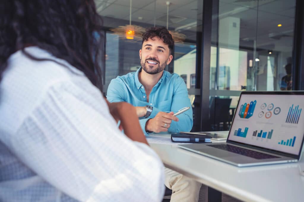 Man in blue shirt discussing charts on a laptop screen with a colleague in an office.