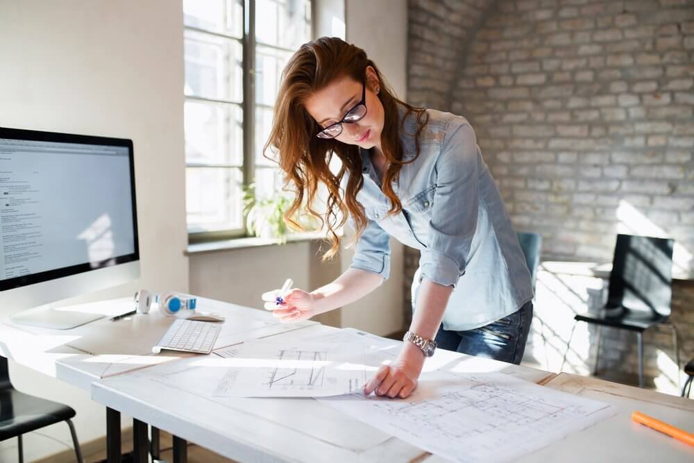 Woman with glasses reviews documents at a desk with a computer in a bright office.