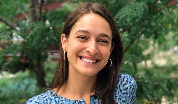 Smiling person with long hair wearing a blue patterned top, standing outdoors with greenery in the background.