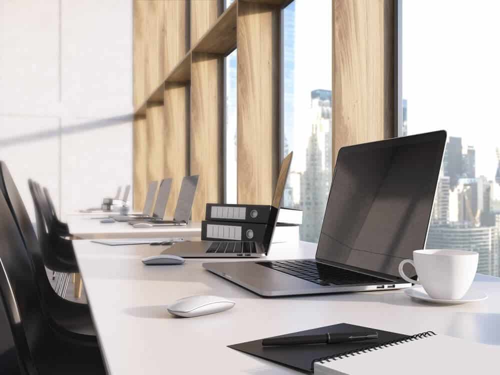 Modern office desk with open laptop, notebooks, pen, and coffee cup; large windows overlook city skyline.
