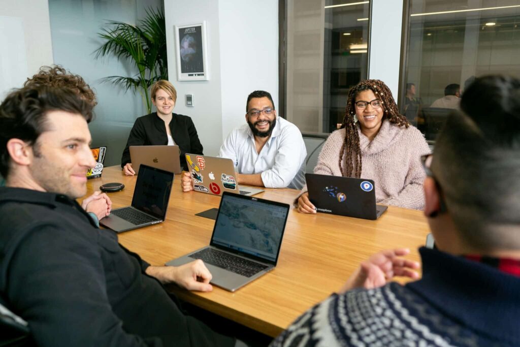 Diverse group of people smiling and talking at a conference table, working on laptops.