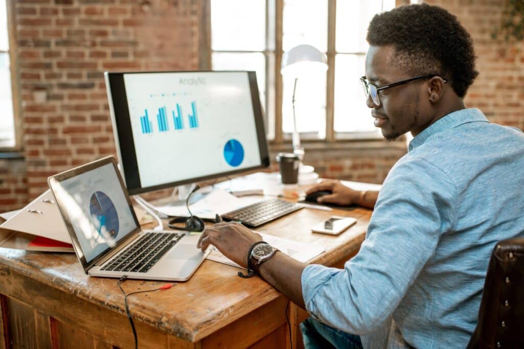 Man working on computers displaying graphs and charts in a brick-walled office.