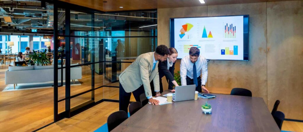 Three people in a conference room reviewing data on a laptop, with charts displayed on a screen.