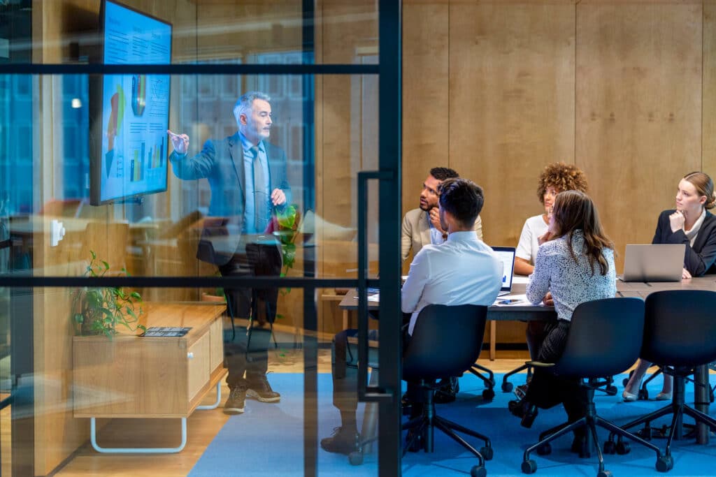 Man presenting graphs on a screen to a seated group in a modern conference room.