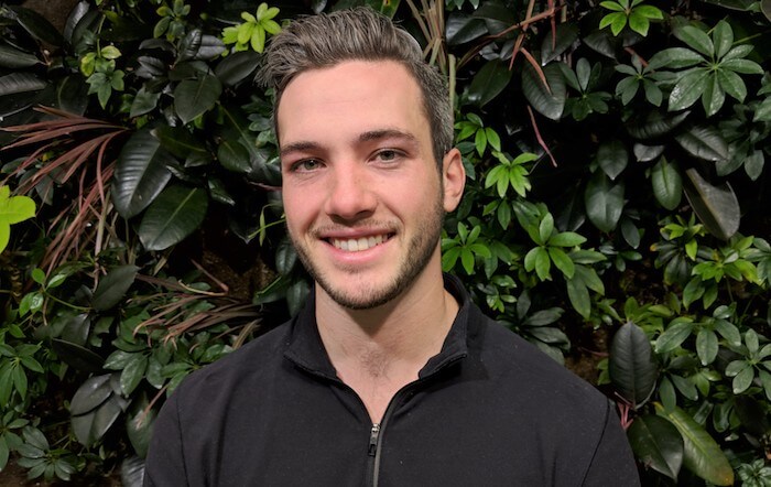 Man in a black shirt smiles in front of a lush green leafy background.