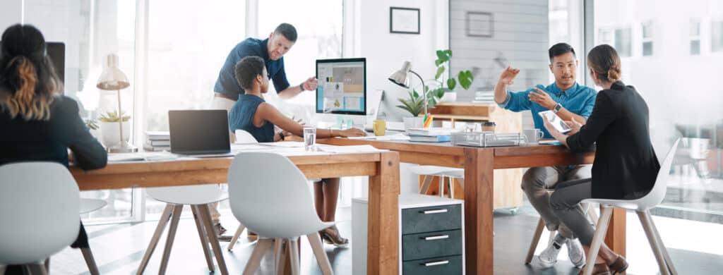 A group of people in a modern office, collaborating around a large table with laptops and papers.