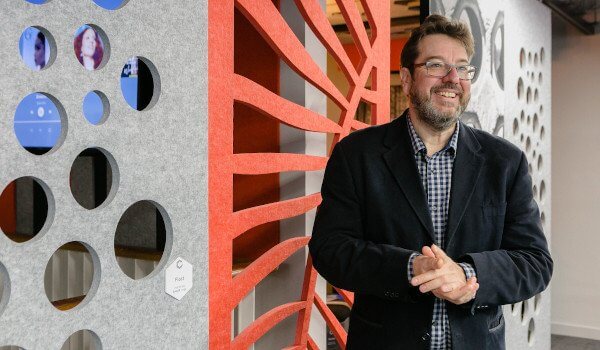 Man in checkered shirt and blazer smiling, standing in front of a decorative wall with circular patterns.