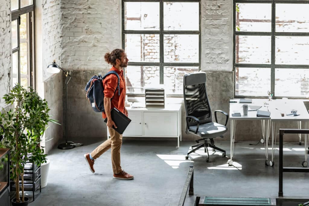A person with a backpack and laptop walks into a modern office with large windows and a desk.