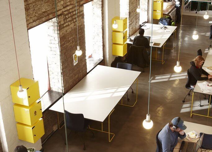 Modern co-working space with people working at long tables, exposed brick wall, and hanging lights.