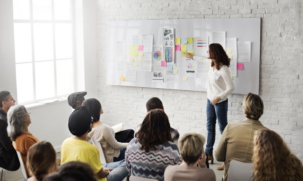 A woman presenting to a diverse group, pointing at a whiteboard covered with papers and sticky notes.