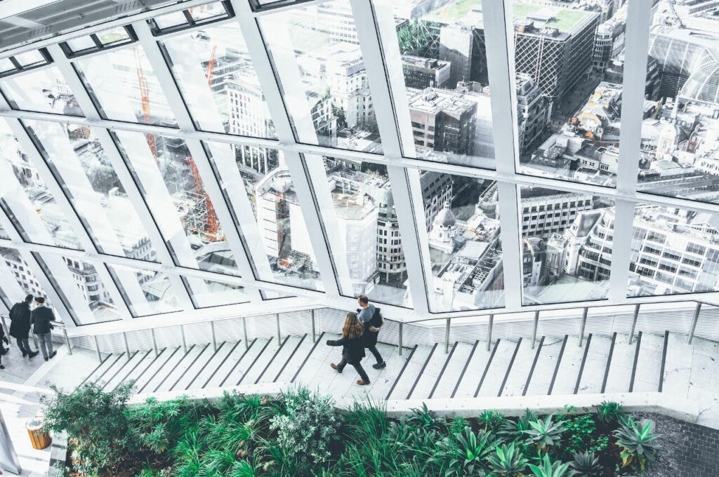 People walking down a staircase inside a large glass building with city views and indoor greenery.