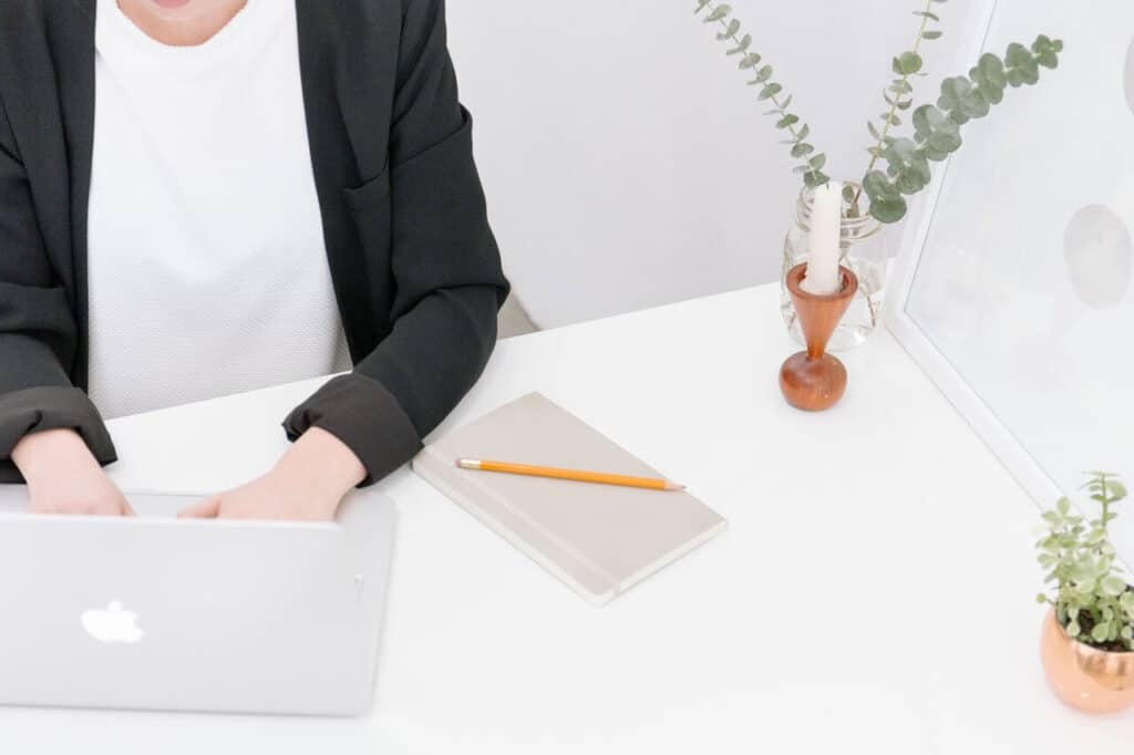 Person typing on a laptop at a white desk with a notebook, pencil, and plants.