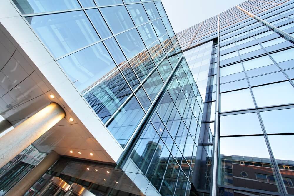Upward view of modern glass skyscraper with reflected sky and clouds, featuring a column near the entrance.