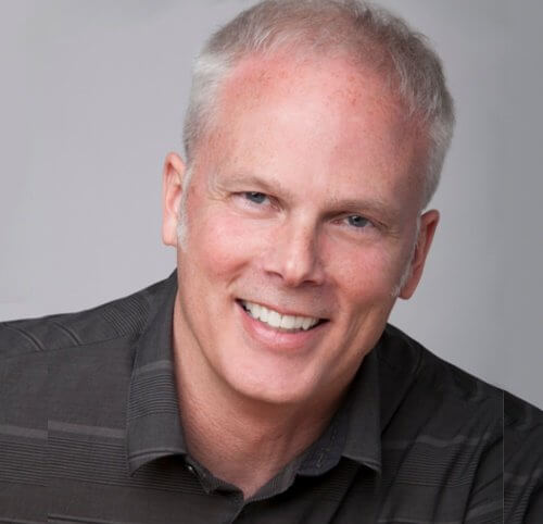Smiling man with short gray hair wearing a dark shirt, looking at the camera against a neutral background.