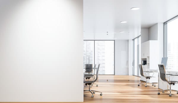 Modern office with empty desks, chairs, large windows, and wooden flooring, divided by a white wall.