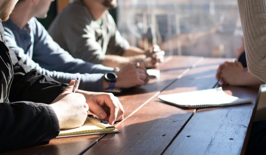 People sitting at a wooden table, taking notes during a meeting or discussion.
