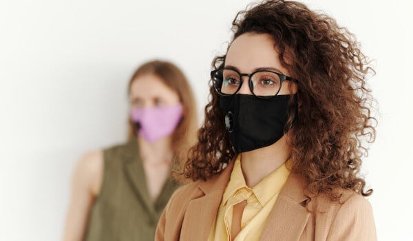 Two people wearing face masks indoors; one in a black mask with glasses, the other in a purple mask in the background.