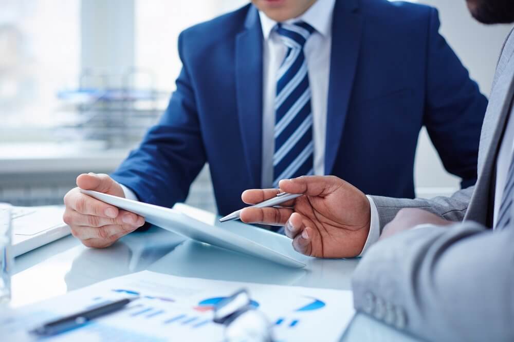 Two people in suits discussing over a tablet at a desk with financial documents.