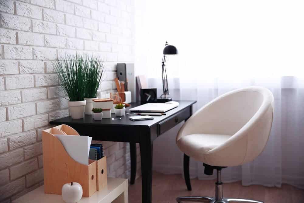 Minimalist home office with a black desk, white chair, desk lamp, potted plants, and a bright window.