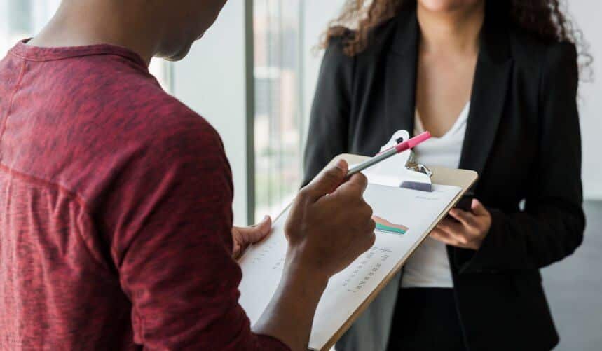 Person holding clipboard with graph, standing in front of another person in business attire.