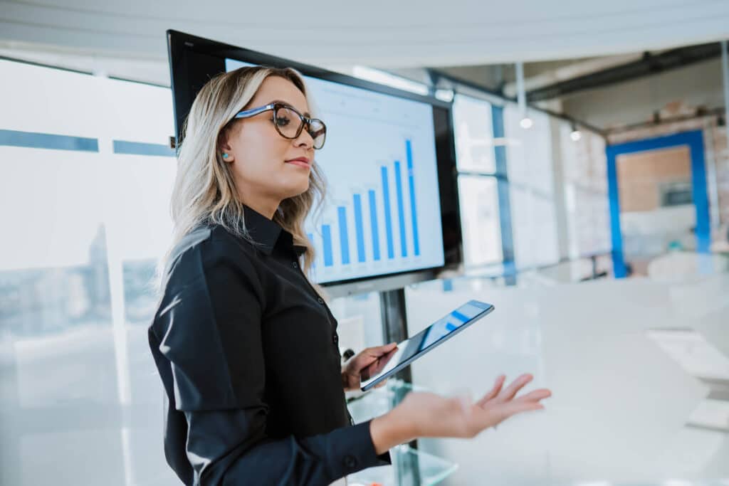 Person presenting with a tablet, standing near a screen displaying a bar graph in an office setting.