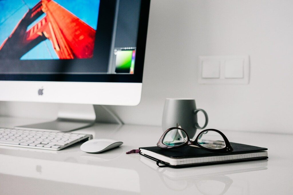 Desk with an iMac, keyboard, mouse, notebook with glasses on top, and a gray mug against a white wall.