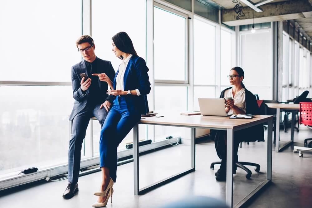 Two professionals discuss something on a phone while sitting on a desk, with another person working at a laptop nearby.