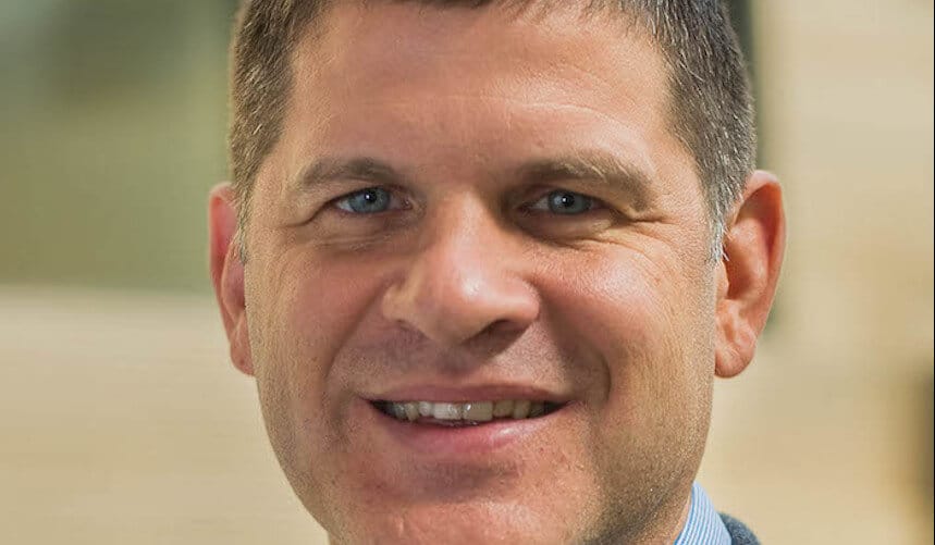 Smiling man with short brown hair, wearing a blue collared shirt, in a blurred indoor setting.