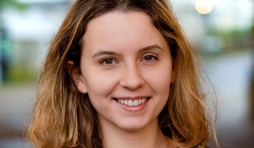 Smiling person with wavy hair, wearing a dark top, stands outside with a blurred background.