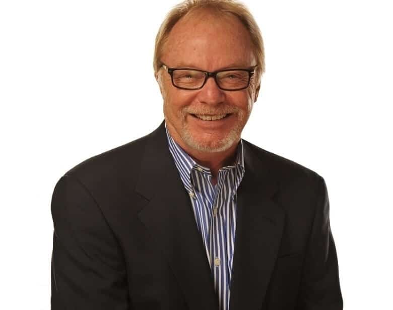 Smiling man with glasses and a beard, wearing a dark blazer over a striped shirt, on a plain white background.
